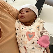 Amina a rejoint le concours — aidez-le/la à gagner de superbes lots ! baby, infant, cute, onesie, heart_pattern, pink_mitten, white_cap, arm, adult, indoor, curious, soft_lighting, serene, person, clothing, fabric, floor, table, cords, relaxation