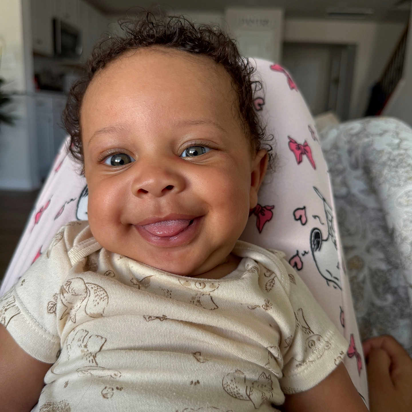 Mazi is registered to the contest to win money with this photo: baby, smiling, tongue_out, curly_hair, blanket, pink, bow_pattern, onesie, infant, indoor, home, kitchen, living_room, person, child, cute, happy, portrait, closeup, playful