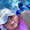 baby, child, hat, pink_clothing, sun_hat, close_up, face, arm, outdoor, blue_background, floral_pattern, curious, selfie, summer, skin, young_child, infant, headwear, casual, daylight