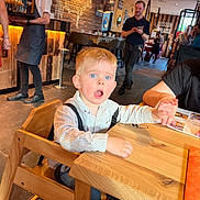 Mael participe au concours pour gagner de l'argent avec cette photo : toddler, child, high_chair, restaurant, table, wooden_furniture, adult_hand, waiter, people, indoor, casual_clothing, blonde_hair, blue_eyes, expression, holding_hand, menu, floor, lighting, brick_wall, dining