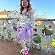 Joy a rejoint le concours — aidez-le/la à gagner de superbes lots ! child, girl, smiling, curly_hair, white_sweatshirt, lilac_skirt, pink_sneakers, balcony, outdoor, daylight, blue_sky, residential_area, playful_pose, hair_bows, concrete_railing, wooden_floor, green_grass, soccer_goal, casual_clothing, portrait
