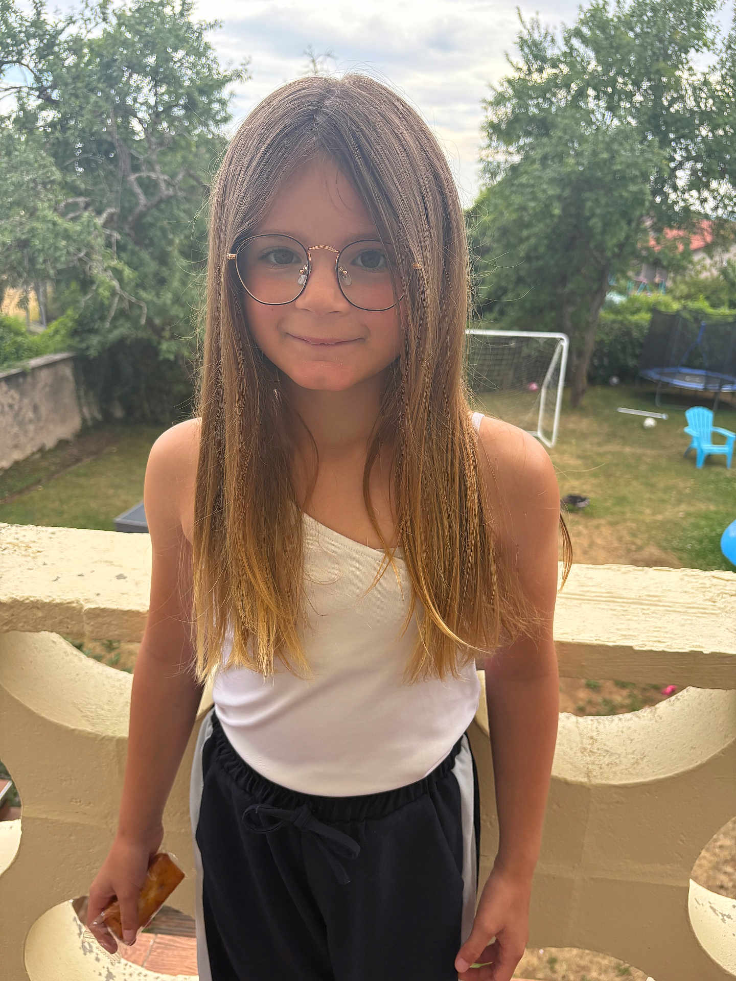 Joy participe au concours pour gagner de l'argent avec cette photo : child, girl, glasses, long_hair, white_top, black_pants, snack, balcony, outdoor, daylight, backyard, soccer_goal, trampoline, grass, trees, smile, casual_clothing, fence, nature, recreation