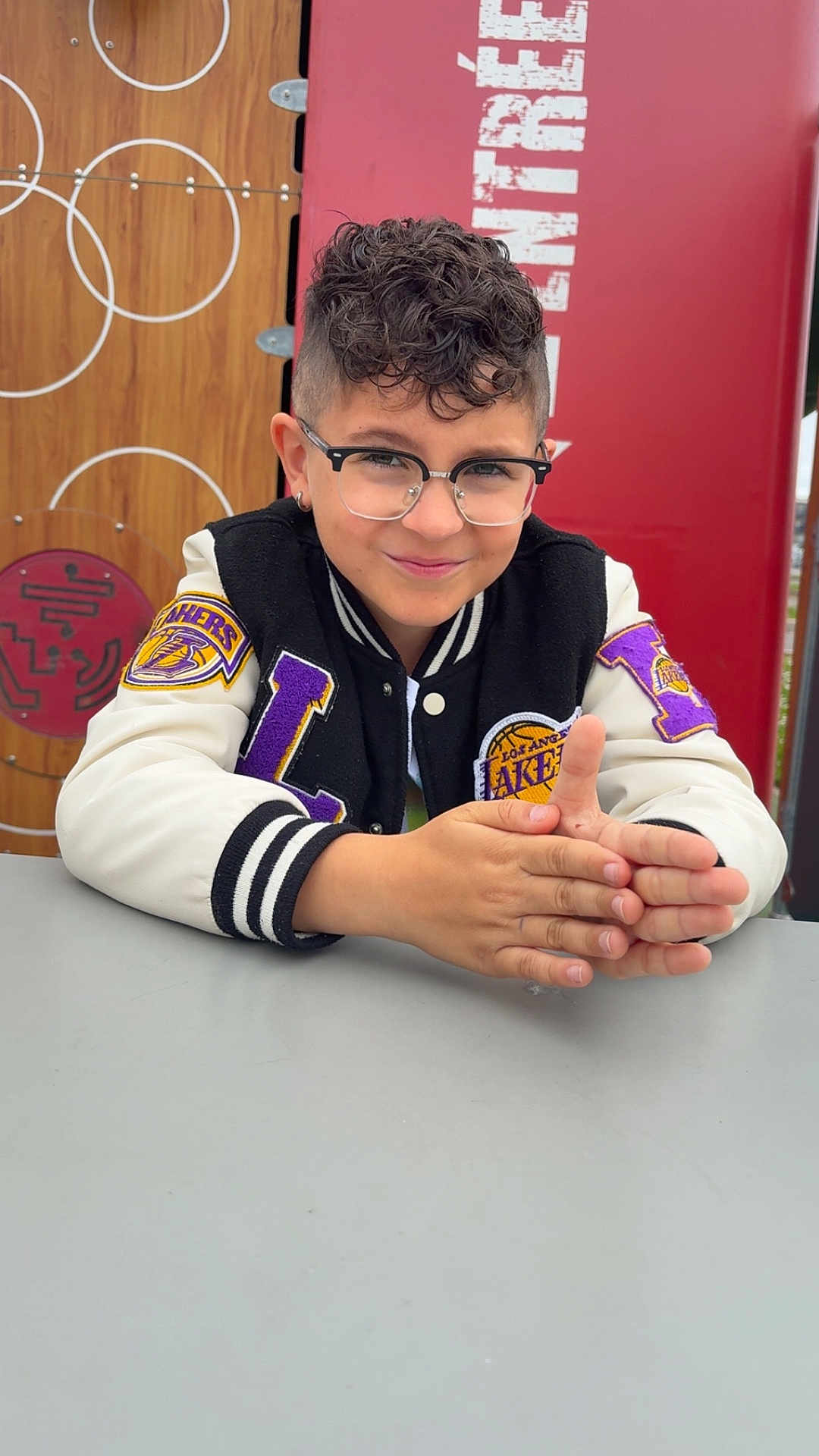 Menzo participe au concours pour gagner de l'argent avec cette photo : child, boy, glasses, curly_hair, jacket, los_angeles_lakers, table, smile, portrait, casual, indoor, person, fashion, hands, seated, young, confident, background, red, wood