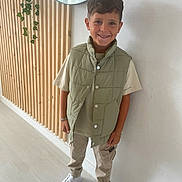 Menzo participe au concours pour gagner de l'argent avec cette photo : child, boy, smile, standing, indoor, casual_clothing, puffer_vest, beige_pants, white_sneakers, bracelet, wooden_wall, mirror, green_plant, modern_interior, happy, portrait, fashion, short_hair, light, floor