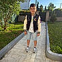 child, boy, glasses, jacket, shorts, sneakers, pathway, garden, grass, tree, shrub, sunlight, daytime, casual_clothing, outdoor, smile, standing, greenery, fence, lamp_post