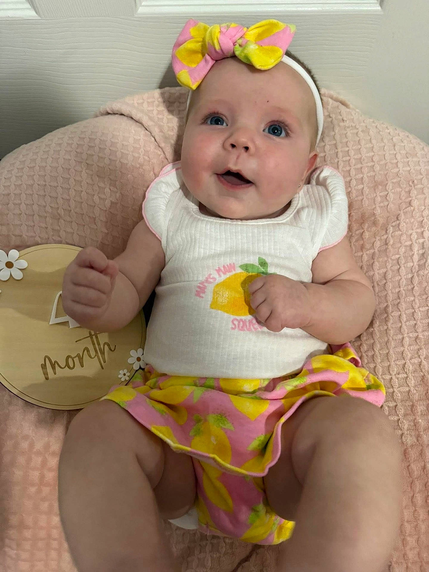 4_months, baby, blue_eyes, bow, child, clothing, cute, furniture, happy, headband, indoor, infant, lemon_pattern, person, pink, pink_blanket, portrait, sitting, wooden_sign, yellow