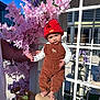 baby, infant, child, red_hat, overalls, corduroy, bicycle, saddle, pink_flowers, purple_flowers, flower_arrangement, window_frame, adult_arm, hand, outdoor, sunlight, portrait, cute, neutral_expression, accessory