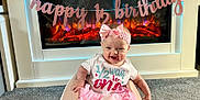 Amelia is registered to the contest to win money with this photo: baby, infant, girl, pink_tutu, headband, smiling, baby_seat, carpet, fireplace, decorations, banner, happy, celebration, indoor, half_birthday, cute, young_child, clothing, home, festive