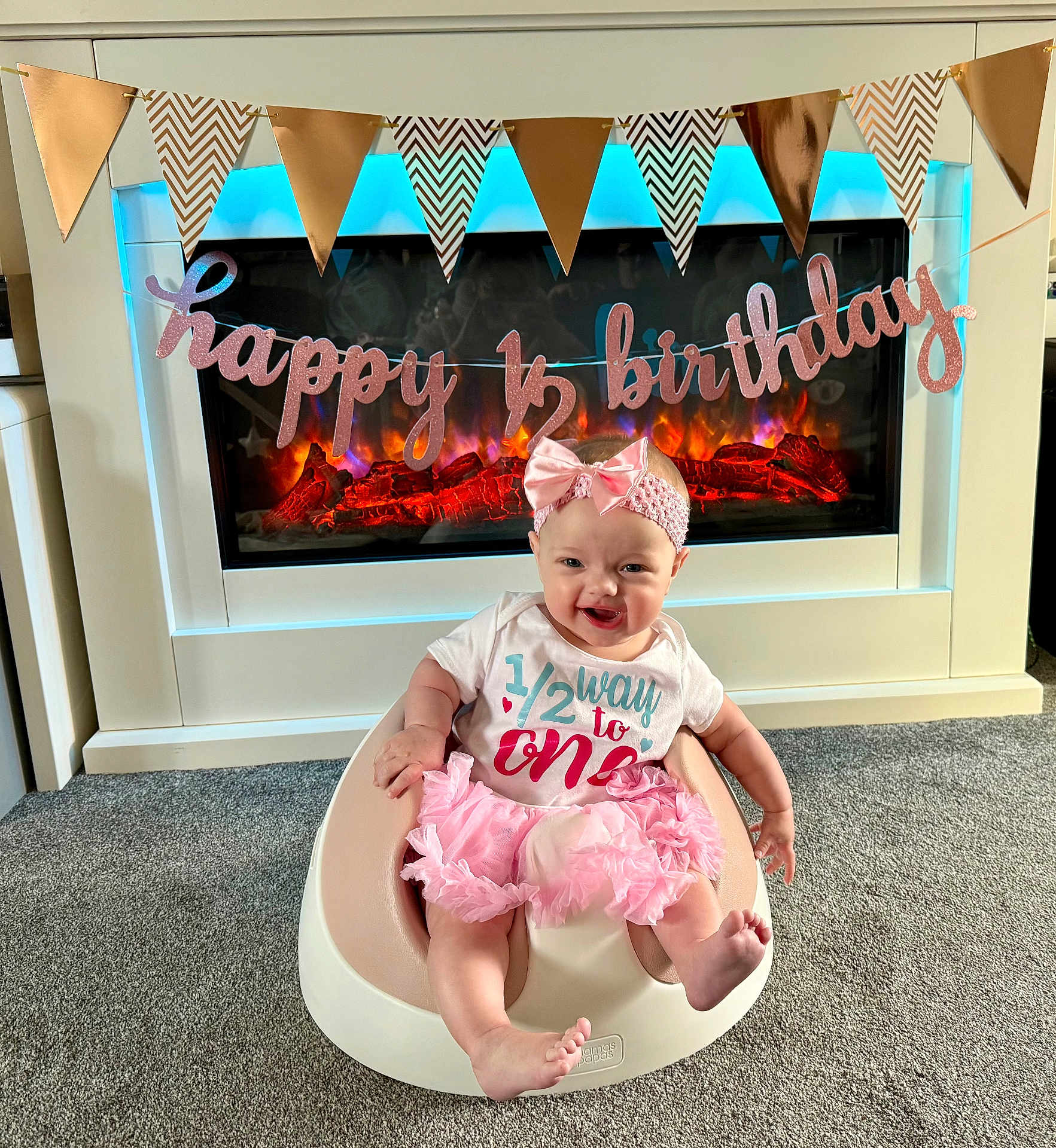 Amelia is registered to the contest to win money with this photo: baby, infant, girl, pink_tutu, headband, smiling, baby_seat, carpet, fireplace, decorations, banner, happy, celebration, indoor, half_birthday, cute, young_child, clothing, home, festive