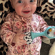 Amelia is registered to the contest to win money with this photo: baby, child, smiling, floral_onesie, rattle, toy, blanket, leopard_print, happy, face, hand, infant, cute, lying_down, indoors, closeup, person, playful, expression, soft