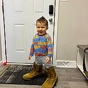 Yasin is registered to the contest to win money with this photo: toddler, child, boots, oversized, indoor, welcome_mat, door, sweater, pants, floor, smiling, standing, person, hair, light_switch, vent, rug, home, playful, funny