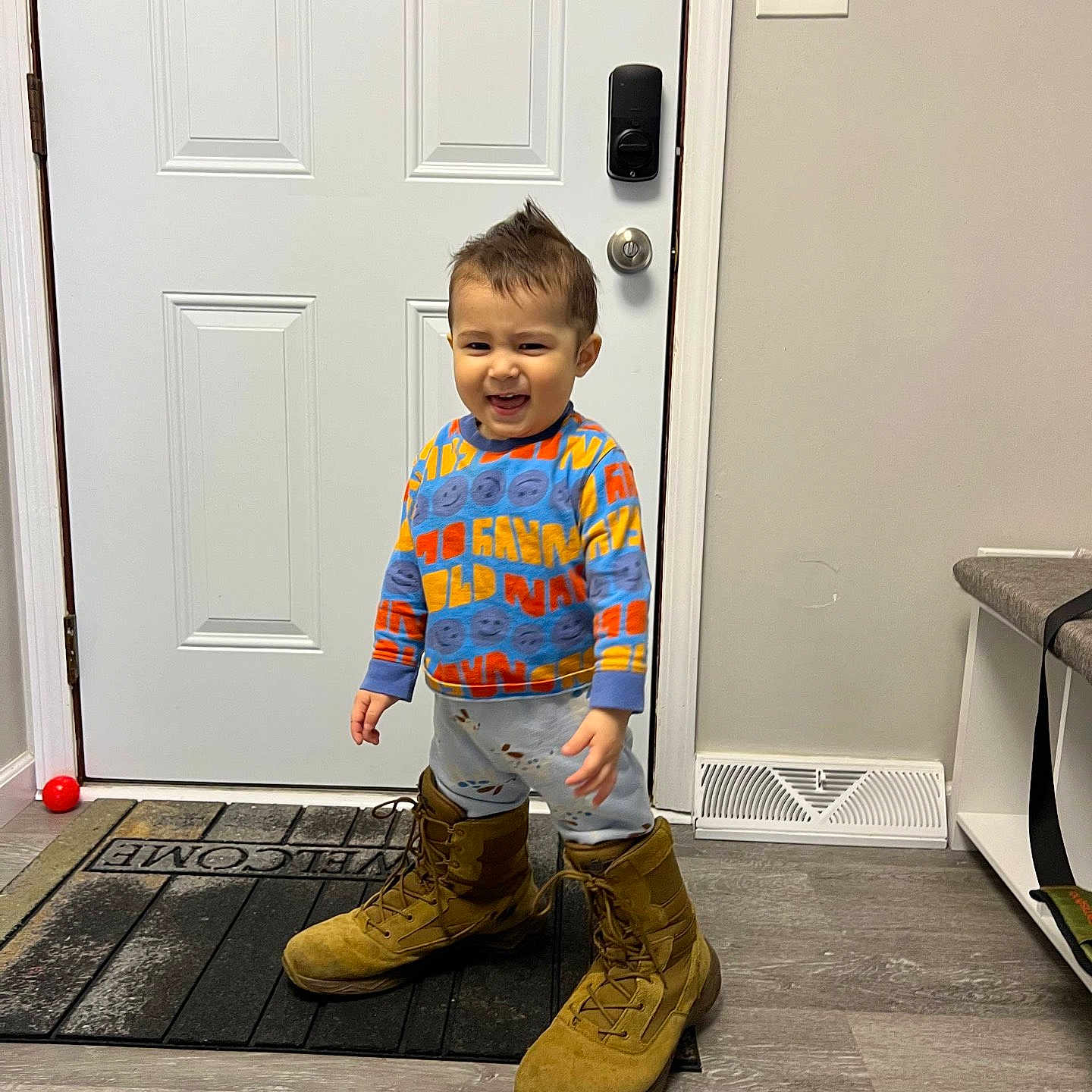 Yasin is registered to the contest to win money with this photo: toddler, child, boots, oversized, indoor, welcome_mat, door, sweater, pants, floor, smiling, standing, person, hair, light_switch, vent, rug, home, playful, funny