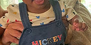 Kyrie is registered to the contest to win money with this photo: baby, child, indoor, person, clothing, mickey_mouse, donald_duck, pluto, overalls, window, greenery, blanket, hand, smiling, cute, tongue_out, playful, young, infant, family