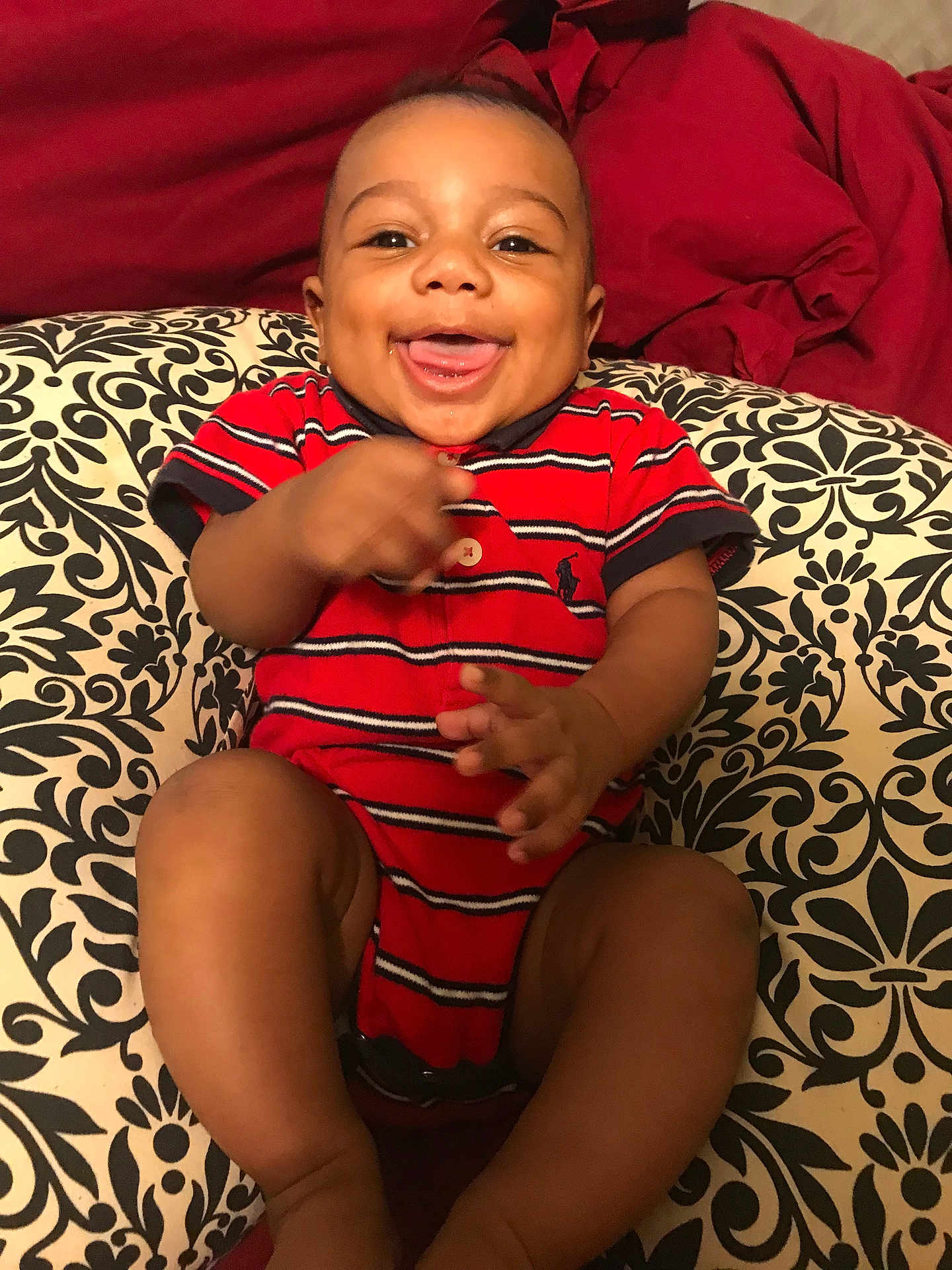 Kyrie joined the competition — help win amazing prizes! baby, child, smiling, happy, tongue_out, red_onesie, striped_clothing, cushion, patterned_cushion, black_and_white, red_blanket, indoor, person, infant, cute, sitting, hands, legs, face, joy