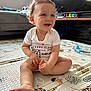 baby, child, smiling, sitting, curly_hair, indoor, playmat, foot, hand, white_onesie, toy, wooden_blocks, television, couch, water_bottle, window, natural_light, floor_mat, living_room, happy