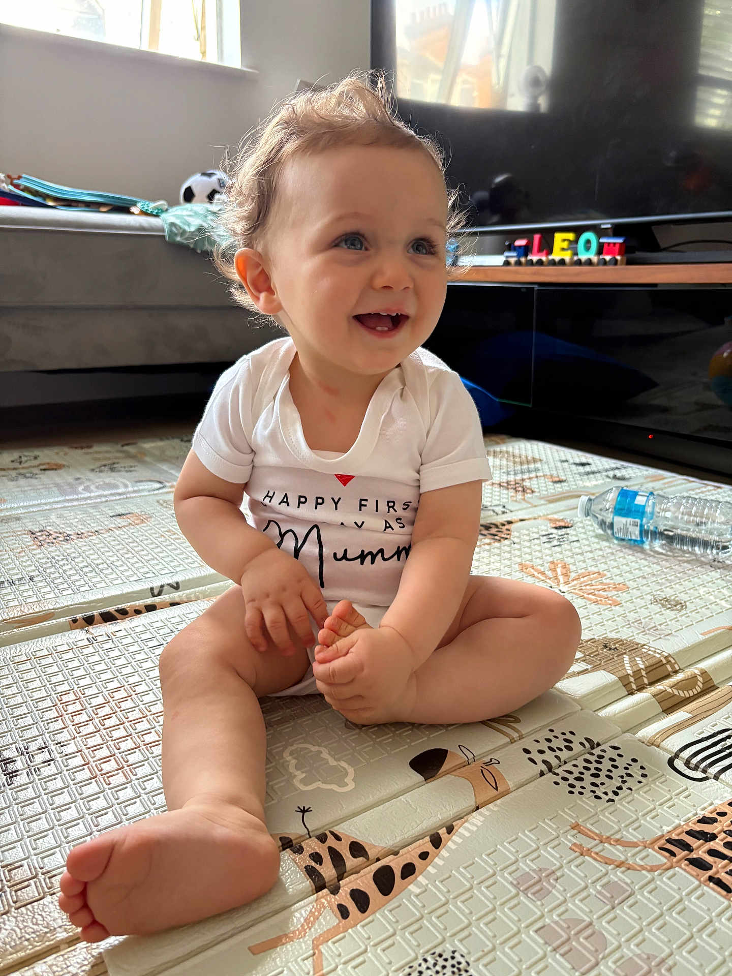 Leo joined the competition — help win amazing prizes! baby, child, smiling, sitting, curly_hair, indoor, playmat, foot, hand, white_onesie, toy, wooden_blocks, television, couch, water_bottle, window, natural_light, floor_mat, living_room, happy