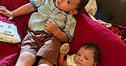 Moses is registered to the contest to win money with this photo: child, baby, toddler, quilt, red, blanket, clothing, shirt, shorts, mittens, socks, shoes, couch, bedroom, window_blinds, person, indoor, cute, family, relaxing