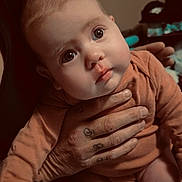 Willow is registered to the contest to win money with this photo: baby, infant, child, person, hand, tattoo, clothing, orange_outfit, face, eyes, skin, expression, portrait, indoors, warm_lighting, cute, young, human, holding, curious