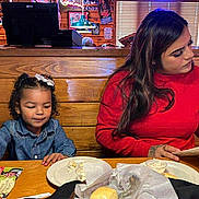 Naomi joined the competition — help win amazing prizes! child, woman, restaurant, menu, table, food, plate, denim_jacket, red_sweater, wooden_table, indoor, people, curtains, bucket, bread, paper, menu_card, casual, seated, waiting