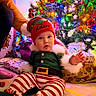 Adan participe au concours pour gagner de l'argent avec cette photo : baby, child, christmas_lights, christmas_tree, colorful, cute, decorations, elf_costume, festive, floor, fur_trim, green_shoes, hand, holiday, indoor, person, presents, red_hat, smiling, striped_pants