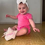 Lynaïa a rejoint le concours — aidez-le/la à gagner de superbes lots ! baby, child, smiling, pink_clothing, headband, bow, indoor, floor, plush_toy, happy, cute, sitting, portrait, person, infant, accessory, jewelry, earrings, wooden_floor, door