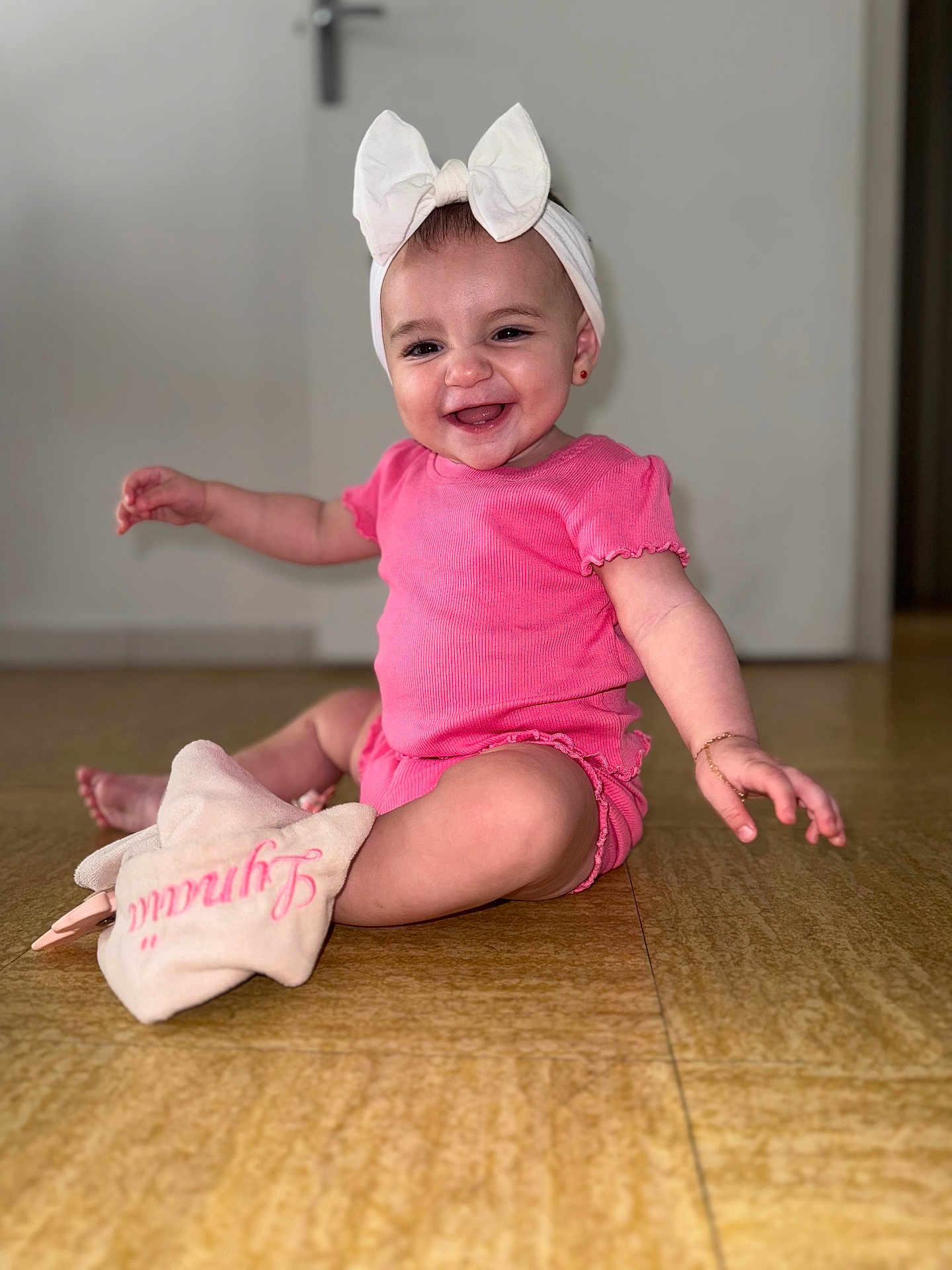 Lynaïa a rejoint le concours — aidez-le/la à gagner de superbes lots ! baby, child, smiling, pink_clothing, headband, bow, indoor, floor, plush_toy, happy, cute, sitting, portrait, person, infant, accessory, jewelry, earrings, wooden_floor, door