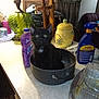 kitten, black_cat, cat, pot, kitchen_counter, lamp, cleaning_supplies, bottle, container, green_eyes, curious, indoor, household_items, ceramic, glass_lamp, purple_bottle, yellow_container, plastic_bottle, curtains, chair