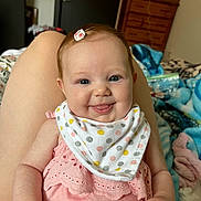 Maddie is registered to the contest to win money with this photo: baby, child, smiling, tongue_out, pink_clothing, bib, hair_clip, indoor, person, infant, happy, face, cute, sitting, legs, bedroom, blanket, furniture, soft_lighting, cozy