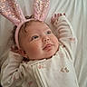 baby, bunny_ears, onesie, infant, cute, playful, lying_down, blue_eyes, sparkly, pink, white_sheet, animal_prints, tongue_out, soft_lighting, child, portrait, indoor, sleepwear, headband, smiling