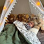 baby, blanket, tent, stuffed_animals, teddy_bears, string_lights, plush_toys, knitted_blanket, smiling, indoor, cozy, child, cushion, fabric, bedroom, decor, person, cute, soft_toys, resting