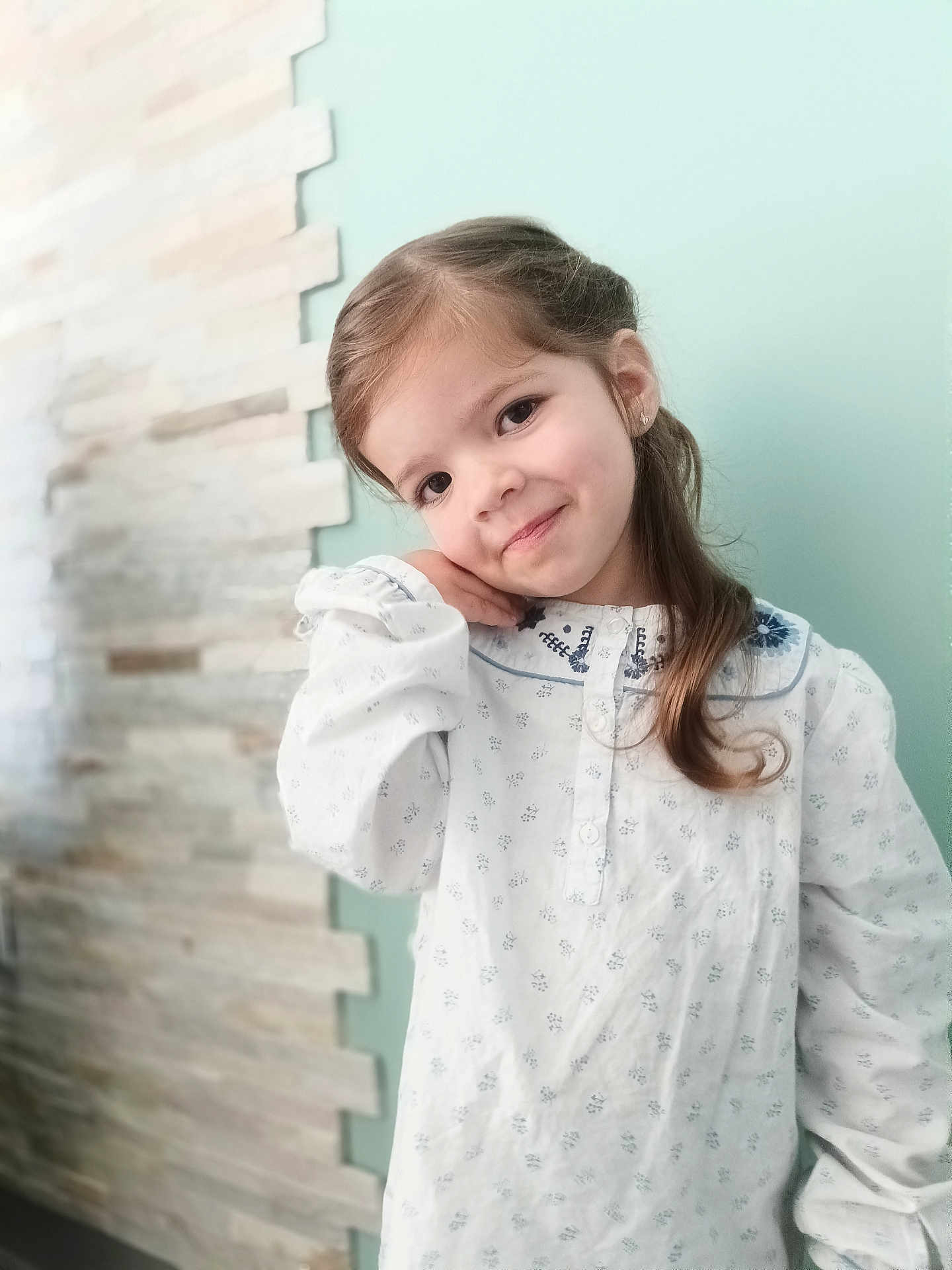Lyana participe au concours pour gagner de l'argent avec cette photo : child, girl, portrait, smile, long_hair, earring, hand_on_cheek, white_dress, patterned_dress, collar, tilted_head, eyes, cute, posing, indoor, pastel_wall, brick_wall, soft_light, young, happy