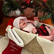 Isla joined the competition — help win amazing prizes! baby, christmas_tree, christmas_lights, ornament, stocking, santa_hat, baby_clothing, knitted_basket, holiday, festive, smiling, indoor, floor, wood_floor, curtain, red, white, green, celebration, cute