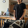 Yoru is registered to the contest to win money with this photo: person, cat, black_cat, couch, cushion, kitchen, water_dispenser, clock, wooden_cabinets, stool, curly_hair, black_tshirt, indoor, home, furniture, appliance, countertop, domestic, relaxed, pet