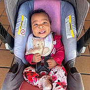 Kaia is registered to the contest to win money with this photo: baby, infant, child, smiling, car_seat, carriage, bib, teddy_bear, floral_clothing, pink_clothing, safety_harness, portrait, indoor, seated, happy, face, hands, feet, wooden_floor, close_up