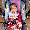 baby, infant, child, smiling, car_seat, carriage, bib, teddy_bear, floral_clothing, pink_clothing, safety_harness, portrait, indoor, seated, happy, face, hands, feet, wooden_floor, close_up