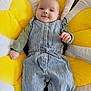 baby, child, smile, face, overalls, striped_clothing, cushion, yellow, flower_shape, indoor, happy, cute, infant, person, portrait, clothing, relaxing, head, hand, comfort