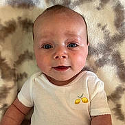 Elliot is registered to the contest to win money with this photo: baby, infant, face, eyes, smile, onesie, clothing, lemon_embroidery, blanket, pattern, skin, arm, cute, child, young, portrait, lying_down, indoor, closeup, baby_garment