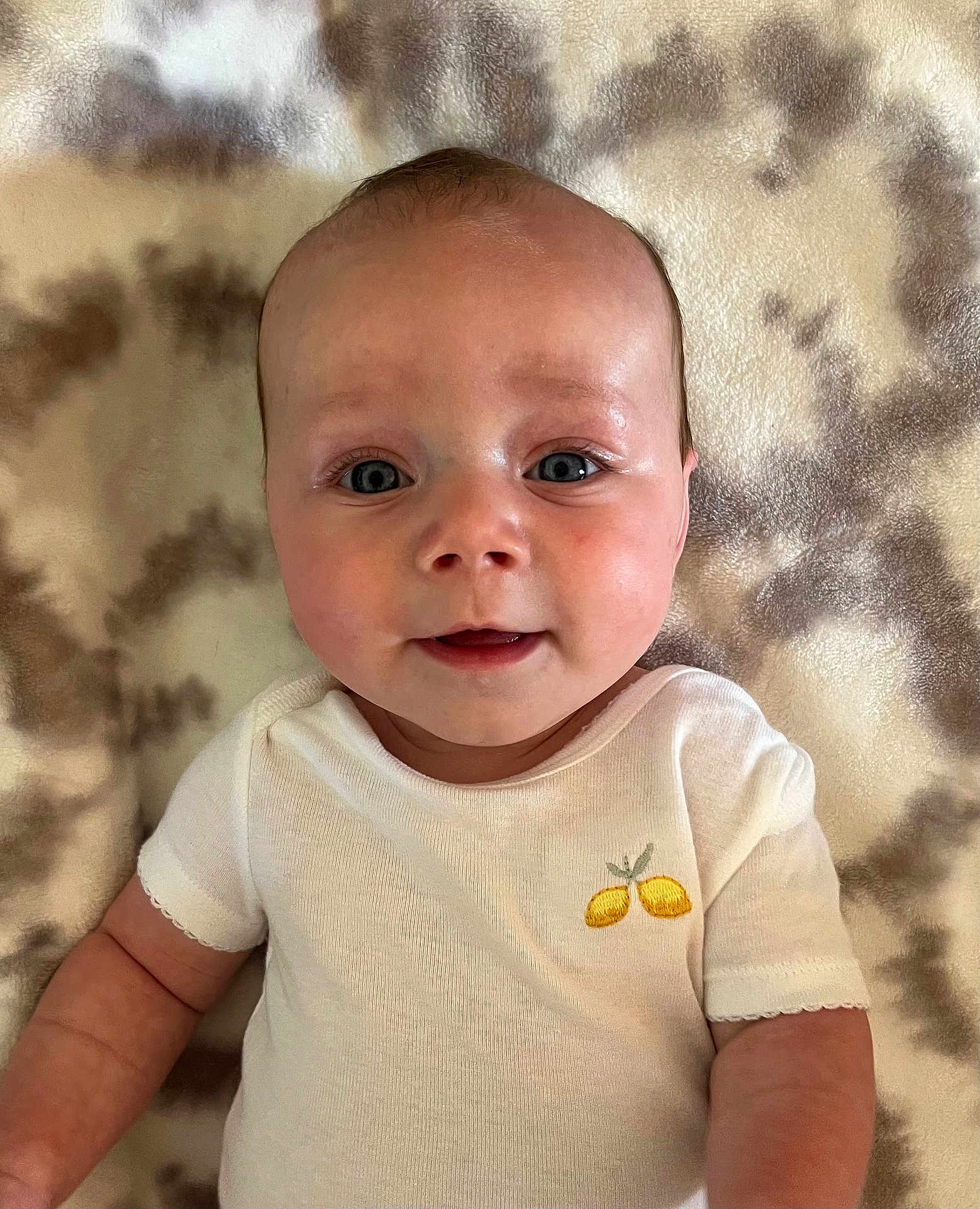 Elliot is registered to the contest to win money with this photo: baby, infant, face, eyes, smile, onesie, clothing, lemon_embroidery, blanket, pattern, skin, arm, cute, child, young, portrait, lying_down, indoor, closeup, baby_garment
