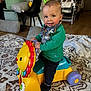toddler, child, toy, ride_on_toy, lion_toy, bandana, green_sweater, black_pants, black_shoes, rug, cushion, chair, high_chair, wooden_floor, indoor, smiling, happy, person, home, playtime