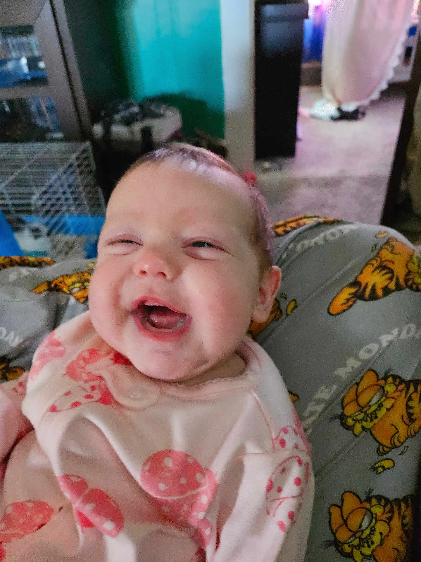 Winters is registered to the contest to win money with this photo: baby, smiling, infant, pink_clothing, polka_dots, garfield, person_holding, indoor, cage, furniture, carpet, face, happy, cute, child, expression, human, casual, home, portrait