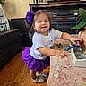 child, toddler, girl, smiling, portrait, indoors, living_room, fireplace, table, tutu, purple_skirt, hair_accessory, flower, white_shirt, silver_shoes, toy, plant, furniture, hardwood_floor, happy