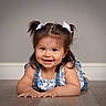 toddler, child, smiling, pigtails, white_bows, denim_overalls, floral_top, indoor, floor, wooden_floor, cute, happy, portrait, baby, person, young_child, casual_clothing, face, head, playful