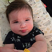 Gavriel is registered to the contest to win money with this photo: baby, infant, face, child, black_shirt, yellow_wristband, hand, cushion, patterned_bedding, animal_print, cloud_print, tousled_hair, skin, person, portrait, indoors, cute, lying_down, expression, soft_lighting