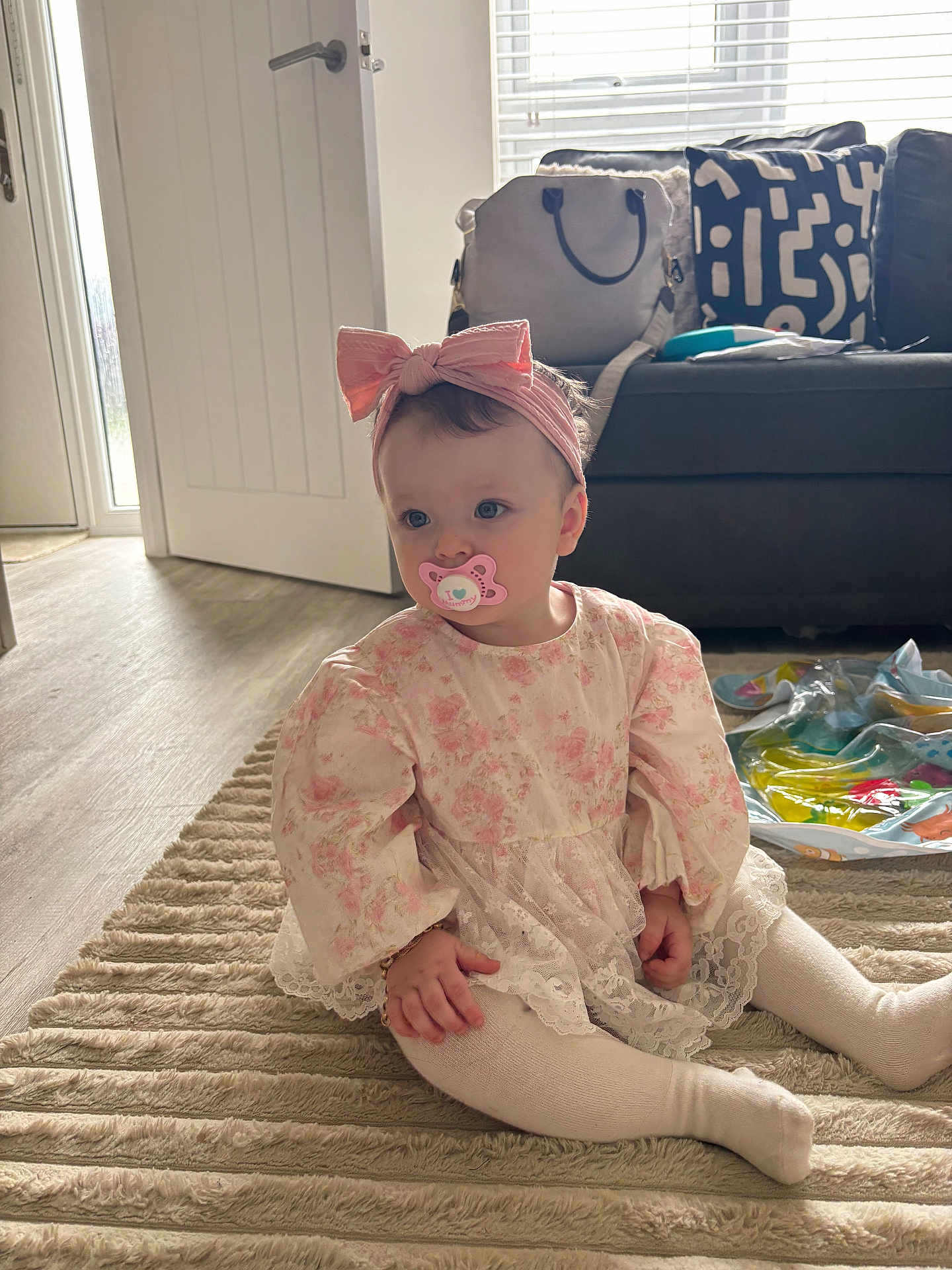 Willow-Rose is registered to the contest to win money with this photo: baby, child, girl, pacifier, headband, bow, dress, floral, tights, rug, floor, living_room, sofa, cushion, bag, window, door, natural_light, toy, indoors