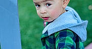 Zaid joined the competition — help win amazing prizes! toddler, child, green_jacket, hood, blue_chair, outdoor, grass, curious, face, hand, expression, young_child, portrait, casual_clothing, daylight, person, short_hair, nature, holding, looking_back