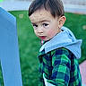 toddler, child, green_jacket, hood, blue_chair, outdoor, grass, curious, face, hand, expression, young_child, portrait, casual_clothing, daylight, person, short_hair, nature, holding, looking_back