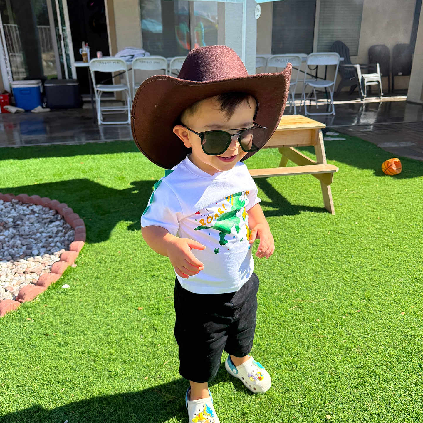 Zaid is registered to the contest to win money with this photo: toddler, child, cowboy_hat, sunglasses, dinosaur_tshirt, grass, backyard, picnic_table, umbrella, shoes, outdoor, sunny, smiling, toy_ball, play, casual_clothing, daylight, shadow, happy, person