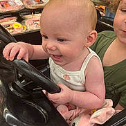 Riley joined the competition — help win amazing prizes! baby, child, toddler, infant, smiling, happy, shopping_cart, supermarket, grocery_store, steering_wheel, plush_toy, bunny, sitting, portrait, face, person, green_shirt, white_onesie, indoor, family