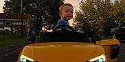 Jude a rejoint le concours — aidez-le/la à gagner de superbes lots ! toddler, child, toy_car, yellow_car, audi_r8_spyder, sunset, street, trees, headlights, car_door, outdoor, residential_area, fence, sky, vehicle, person, side_mirror, curious, evening, road