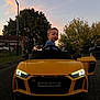 toddler, child, toy_car, yellow_car, audi_r8_spyder, sunset, street, trees, headlights, car_door, outdoor, residential_area, fence, sky, vehicle, person, side_mirror, curious, evening, road